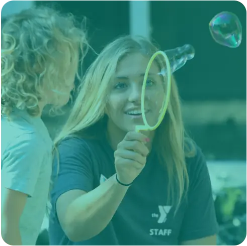 A Y staff member blowing bubbles for a young child outdoors.