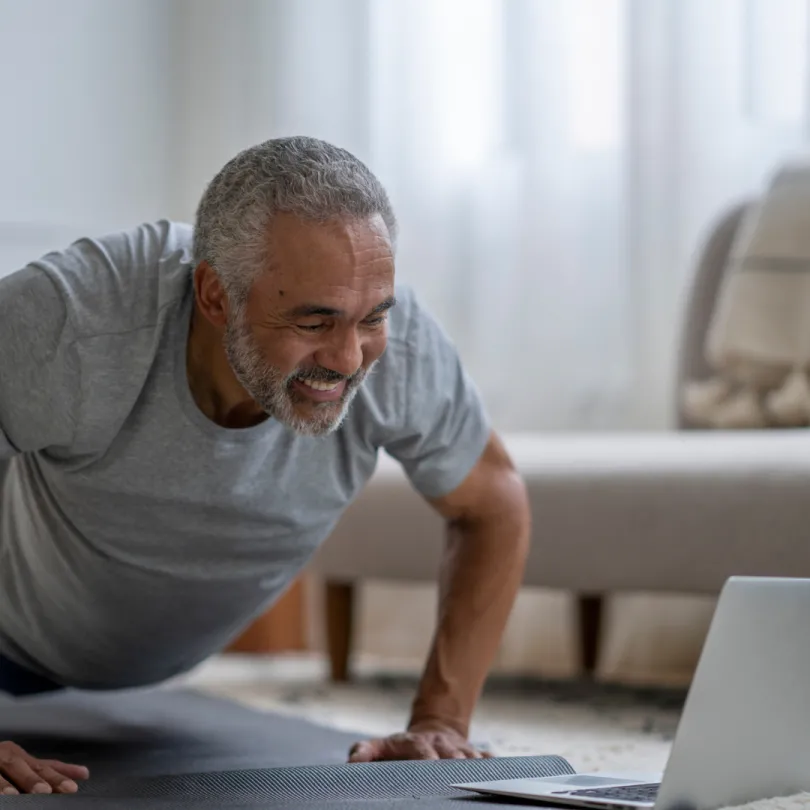An older adult man smiles as he exercises in front of a laptop for a virtual workout routine.