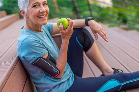 An older woman dressed in activewear smiles at the camera and eats a green apple. She is sitting on stairs outside; a pink yoga mat is laying next to her.