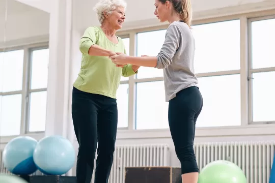 An older woman balancing on a balance ball. Another woman supports her by holding her hands.