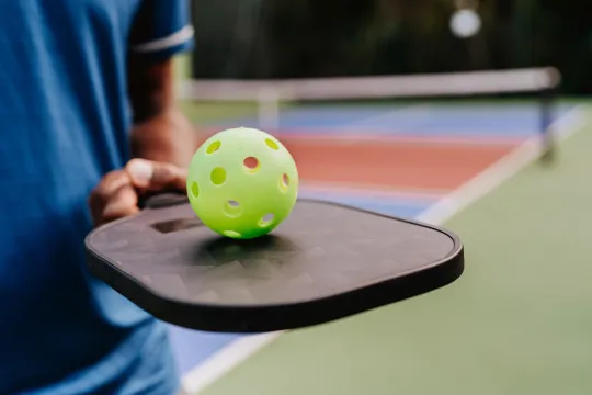 A closeup of someone holding a pickleball paddle and pickleball while on a court.