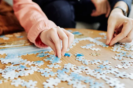 Two people sit at a table and place pieces of a puzzle together.
