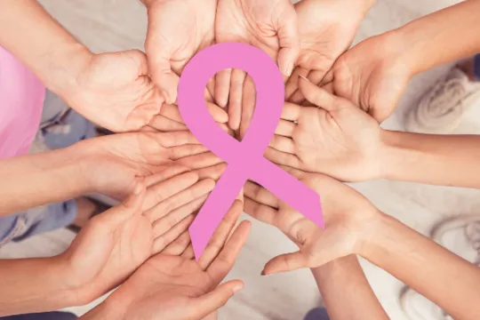 A group of hands hold a pink Breast Cancer Awareness ribbon.