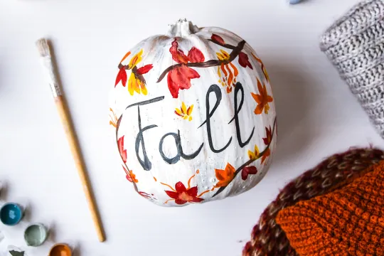 A pumpkin that is painted with autumn leaves and the word "Fall."