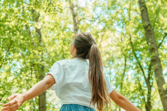 A woman stands in the middle of a forest looking up at the trees.