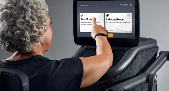 An older woman uses touch screen settings on gym equipment.