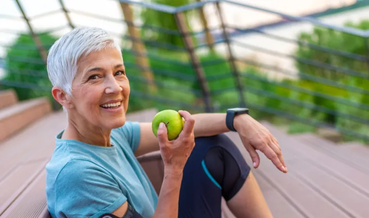 An older woman dressed in activewear smiles at the camera and eats a green apple. She is sitting on stairs outside; a pink yoga mat is laying next to her.