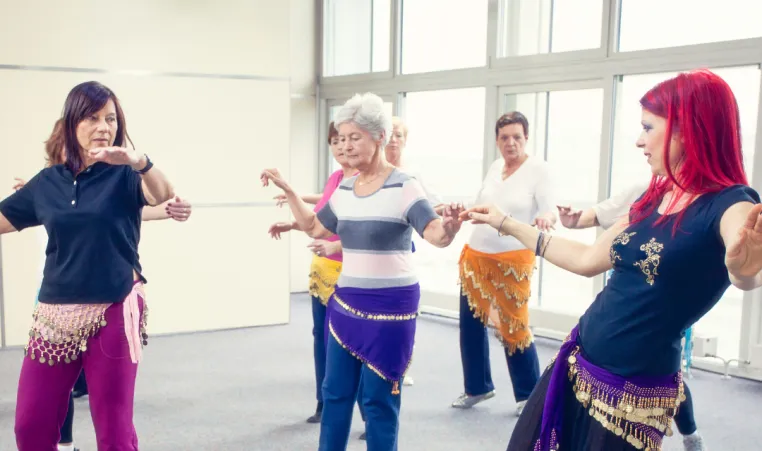 A group of women dance in a belly dancing class. The women are watching the instructor at the front of the group. They are wearing scarves wrapped around their hips.
