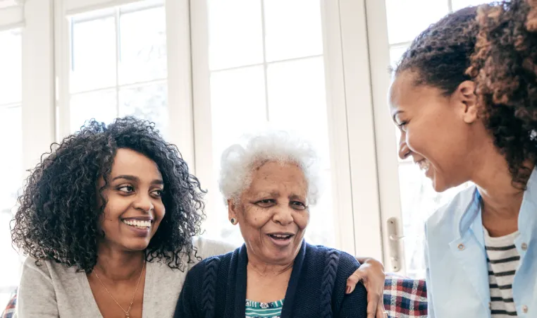 A grandmother sits on the couch with her two grown granddaughters sitting next to her on either side. One granddaught has her arm around her grandmothers' shoulder. They are all smiling and talking with each other.