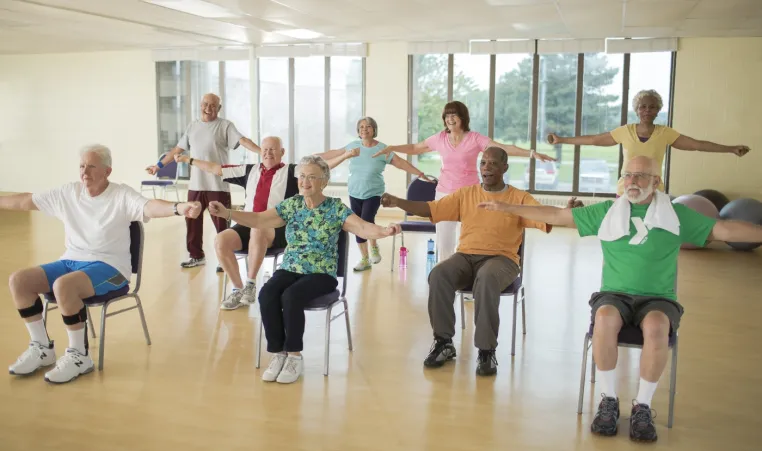 Several people of various race and gender sit in chairs in a workout studio space with their arms extended to each side as if doing arm lifts. 