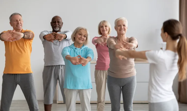 Group of people following instructions from a yoga instructor who appears in the front right corner of photo, stretching hands and arms forward.