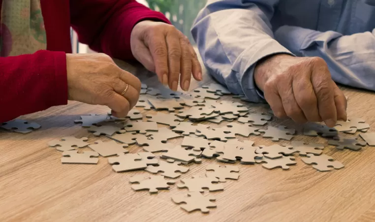 Two elderly people sit next to each other at a table. There are puzzle pieces on the table.