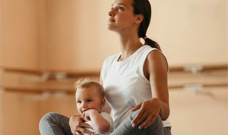 A woman sits on a yoga mat with her baby sitting in her lap.