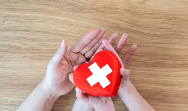 Two pairs of hands hold a red heart shape with a first aid symbol on it.