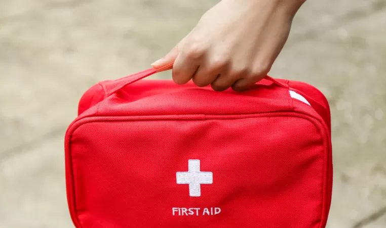 A hand carries a red bag that says "First Aid" below a white cross symbol.