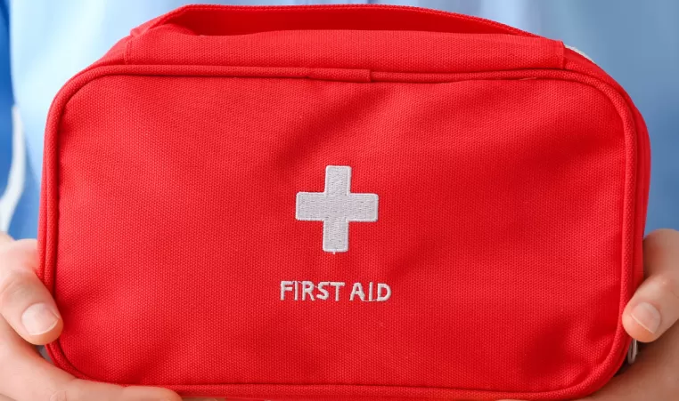 Two hands hold a red bag that says "First Aid" below a white cross symbol.
