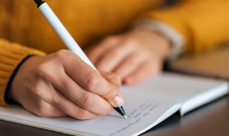 A close up of two hands. The person is holding a pen in one hand and writing in a notebook. They are wearing a yellow sweater.