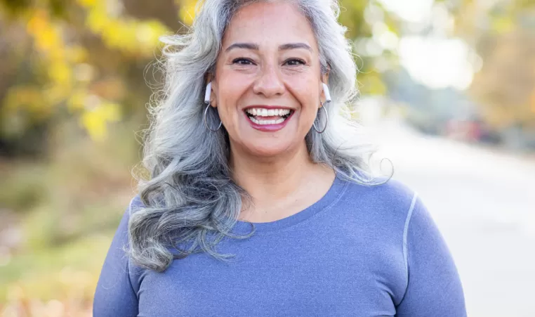 An older woman smiles at the camera as she prepares to start jogging. She is outside and is wearing active wear.