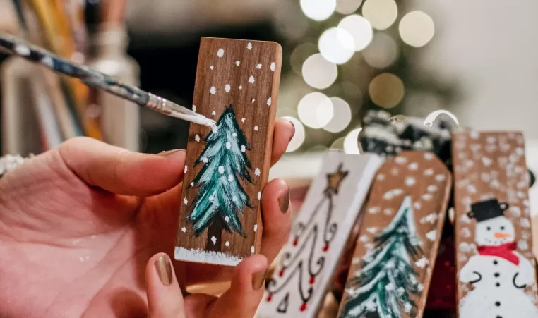 A closeup of a pair of hands painting a Christmas tree on a piece of wood. There are other holiday pieces in the background.