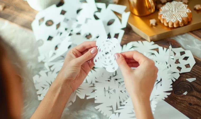 A pair of hands hold a paper snowflake. There are craft supplies around them.