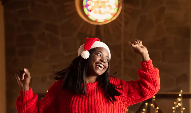 A young woman laughs as she dances indoors. She is wearing a red sweater and a Santa hat. There are string Christmas lights in the background.