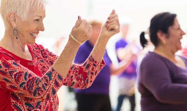 An older woman smiles as she line dances with others in a dance class.