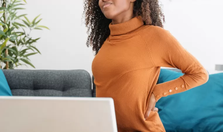 A woman sits on a couch in front of a computer. She is holding her lower back.