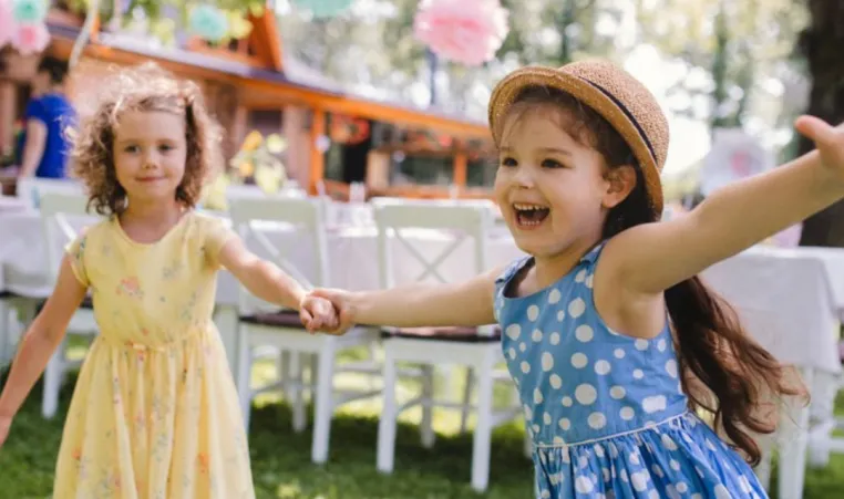 Two girls wearing spring dresses and hats smiles and dance outside in a garden.