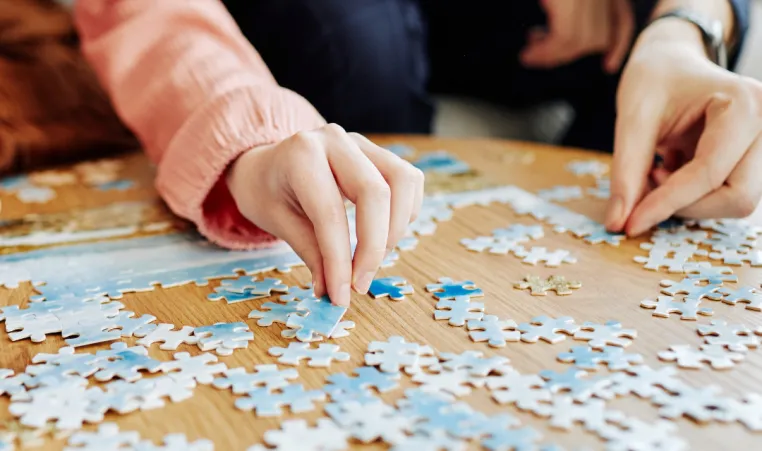 Two people sit at a table and place pieces of a puzzle together.