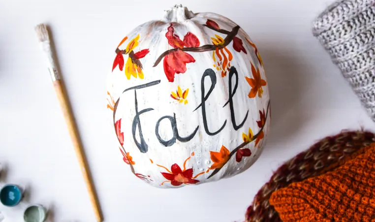 A pumpkin that is painted with autumn leaves and the word "Fall."