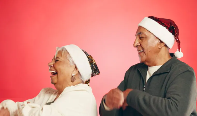 A senior couple dance together during the holidays. They are both wearing Santa hats.