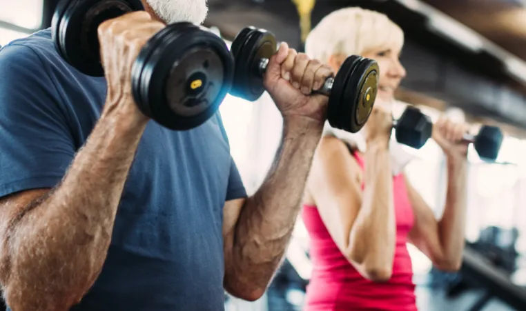Two older adults lift weights inside of a gym.