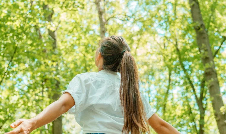 A woman stands in the middle of a forest looking up at the trees.
