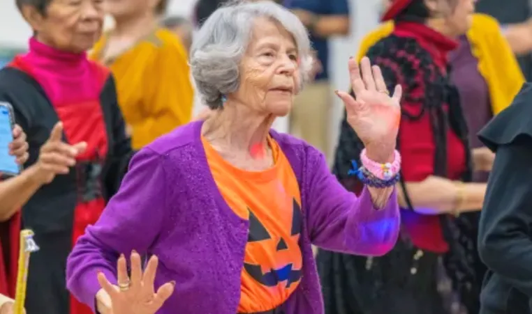 A senior woman wearing a pumpkin shirt dances at a Halloween party.