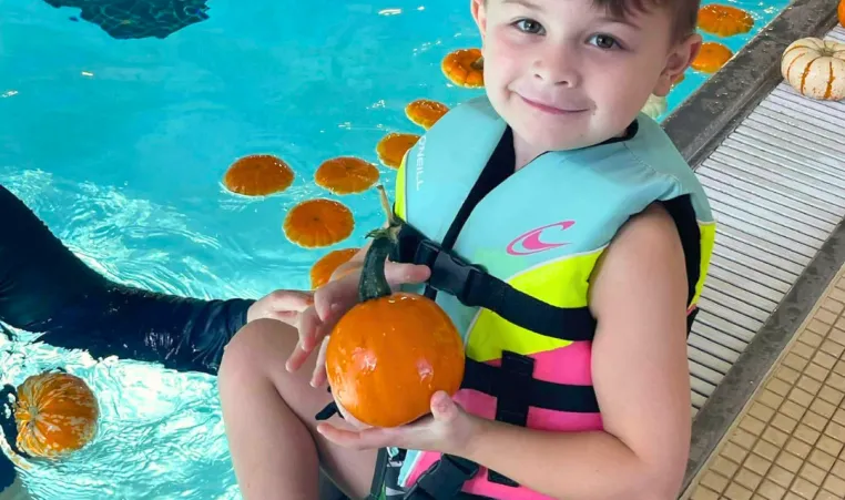 A smiling boy sits next to a pool while holding a small pumpkin.